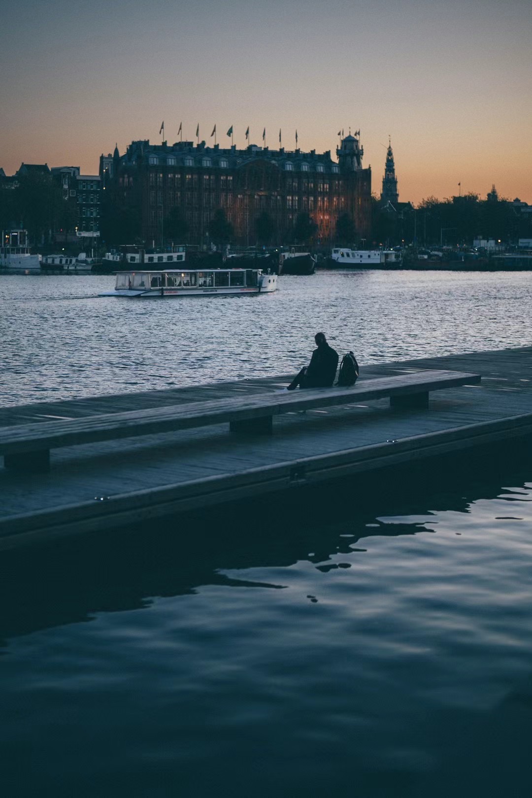 Silhouette seated on a pier in Amsterdam at sunset with boats passing.